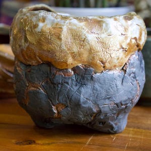 May include: A small, hand-built ceramic planter with a thick, uneven rim featuring a glossy, cream-colored glaze. The lower portion of the vessel has a dark, charcoal-gray textured surface with visible cracks revealing a reddish-brown clay base.