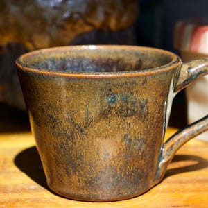 May include: A ceramic mug with a mottled brown and dark bronze glaze. The mug features a wide, slightly flared rim, a rounded body, and a curved handle on the right side. The surface has a textured, earthy appearance with subtle variations in color.