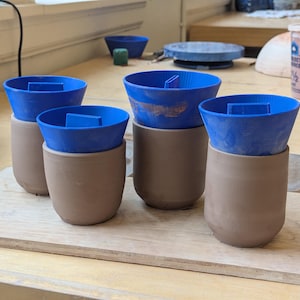 May include: Four unfinished ceramic mugs with blue plastic inserts, arranged on a wooden board. The mugs are a light brown color, and the inserts are a bright blue. The scene suggests a pottery studio or workshop setting.