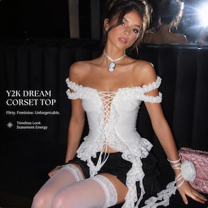 May include: A white off-the-shoulder corset top featuring lace trim, a front lace-up closure, and ruffled sleeves. The garment is paired with black shorts and white lace-trimmed thigh-high stockings. Text on the image reads Y2K DREAM CORSET TOP.