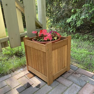 May include: A square wooden planter box with a natural wood finish, filled with vibrant pink and red flowers in a brown pot. The planter is outdoors, sitting on a brick patio.