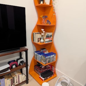 May include: A tall, bright orange corner bookshelf with a distinct wavy, serpentine silhouette. The unit features five open shelves holding various items including potted plants, a glass vase with flowers, books, video games, and board games.