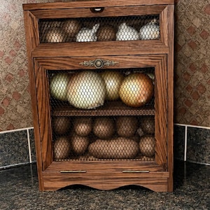 May include: A wooden vegetable storage bin with two tiers and a wire mesh front. The top tier holds onions and garlic, while the bottom tier contains potatoes. The unit features a dark wood finish, a decorative metal accent, and two small handles.