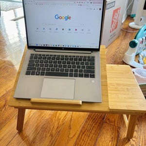 May include: A silver laptop with a black keyboard and trackpad is open on a light brown bamboo desk tray. The screen displays the Google search engine homepage with a search bar, various browser icons, and a taskbar at the bottom of the display.
