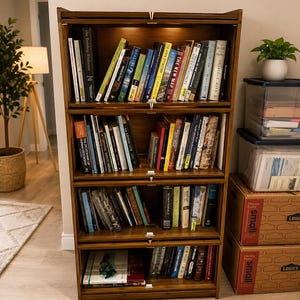 May include: A four-tier wooden bookcase with glass-front doors on each shelf. The unit is filled with various books arranged vertically. Each shelf features a small brass-colored handle at the center of the top edge for opening the glass panels.