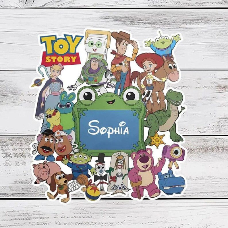 Puede incluir: Una pegatina decorativa con varios personajes de Toy Story que rodean un marco central con forma de rana verde con el nombre Sophia impreso en blanco. Los personajes incluyen a Woody, Buzz Lightyear, Jessie, Rex y otros en un collage.