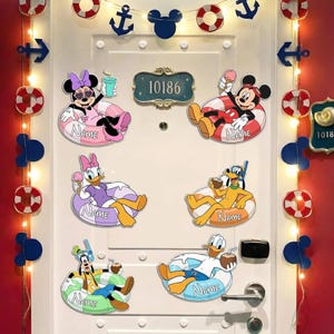 May include: A white door decorated with six Disney character cutouts, each featuring a character in a pool float with the word Name printed on it. Characters include Minnie, Mickey, Daisy, Pluto, Goofy, and Donald. A string of lights with anchors and Mickey heads frames the door.