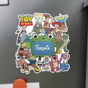 Puede incluir: Un im&aacute;n decorativo con un collage de personajes de Toy Story que rodean un marco central verde con forma de rana y el nombre Sophia impreso en letra cursiva blanca. Los personajes incluyen a Woody, Buzz Lightyear, Jessie y Bullseye.