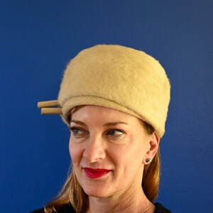 May include: A woman wearing a cream-colored felt hat with a wide brim and a small, round crown. The hat has a simple design and is accented with a thin, brown ribbon.