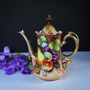 May include: A decorative ceramic teapot with a gold handle and spout. The teapot is painted with a colorful fruit design, including grapes, apples, and pears. The background is a light tan color with a dark brown top and lid.