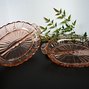 May include: Two vintage pink glass divided serving dishes with scalloped edges and ornate designs. The dishes have handles on either side.