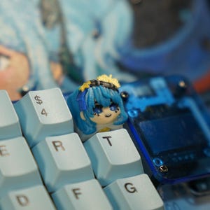 May include: A custom keyboard with light blue keycaps. A keycap featuring a blue-haired anime-style character is on the "T" key. The keyboard also has a clear blue accent piece. The background shows a blurred image of a similar character.