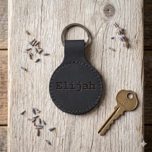 May include: A black leather keychain with the name "Elijah" embossed on a circular tag. A metal key and dried lavender sprigs are next to the keychain on a wooden surface.
