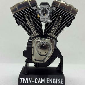 May include: A detailed scale model of a V-twin engine featuring metallic silver and black components, mounted on a black display base with the text "TWIN-CAM ENGINE" and "made by Scale Model Choppers" printed in white on the front of the stand.