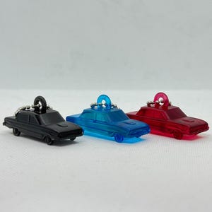 以下が含まれることがあります： 黒、青、赤の3つのミニカーキーホルダー。各車の上部には銀色のリングが付いています。車は小さく、クラシックなセダンのデザインで、半透明の素材で作られています。