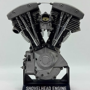 May include: A detailed model of a Shovelhead engine, in shades of grey and silver. The engine features intricate components, including cylinders, carburetors, and cooling fins. The model is mounted on a black base with the text "SHOVELHEAD ENGINE" and "made by Scale Model Choppers".