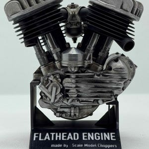 May include: A detailed model of a flathead engine, mainly silver and black. The engine features ribbed cylinders and intricate mechanical components. The base displays the text "FLATHEAD ENGINE" and "made by : Scale Model Choppers".