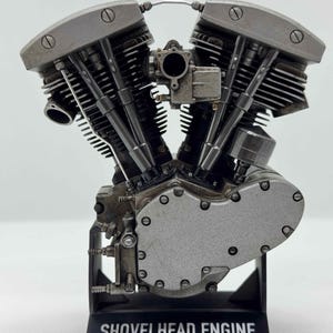 May include: A detailed model of a Shovelhead engine, made by Scale Model Choppers. The engine is primarily silver with visible mechanical components, including cylinders, valves, and a carburetor. The model sits on a black base with the engine's name.