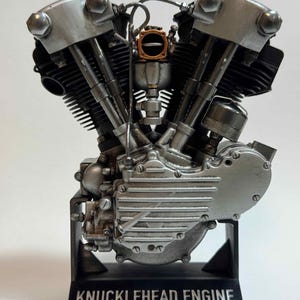 Puede incluir: Un modelo a escala detallado de un motor de motocicleta en V con componentes en plata metálica y negro. El motor presenta una cubierta lateral acanalada prominente y un carburador central. Está montado sobre una base negra con la etiqueta KNUCKLEHEAD ENGINE made by Scale Model Choppers.