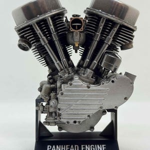 Könnte beinhalten: Detailliertes silbernes Modell eines Panhead-Motors, hergestellt von Scale Model Choppers. Der Motor verfügt über Zylinder, Kühlrippen und einen Vergaser. Montiert auf einem schwarzen Ständer mit der Aufschrift 'PANHEAD ENGINE'.