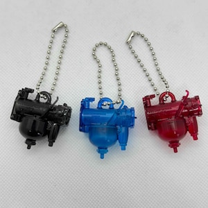 以下が含まれることがあります： 機械部品の形をした、3つのミニチュアキーホルダー。黒、青、赤の3色です。各キーホルダーには、シルバー色のチェーンと留め金が付いています。