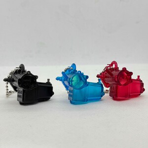 以下が含まれることがあります： 機械装置の形をした、3つのミニチュア透明キーホルダー。1つは黒、1つは青、1つは赤です。各キーホルダーには、小さな金属チェーンとリングが付いています。