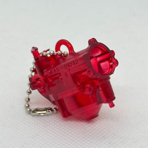 以下が含まれることがあります： 透明な赤いミニチュアキャブレターキーホルダー。キーホルダーはシルバーのボールチェーンに付いています。「SAS-SUPER」の文字がキャブレターの側面に表示されています。