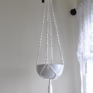 May include: White macrame plant hanger with a white ceramic pot hanging from a black hook on a white wall.