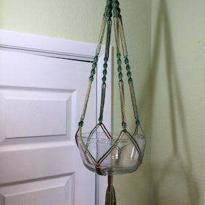 May include: A green and gold macrame plant hanger with a glass planter. The hanger is made with a knot pattern and has a tassel at the bottom.