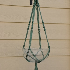 May include: A green macrame plant hanger with a glass bowl and a tassel. The hanger is hanging from a hook on a light beige wall.