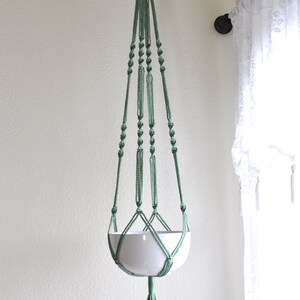 May include: A green macrame plant hanger with a white ceramic pot. The hanger is hanging from a hook on a white wall.