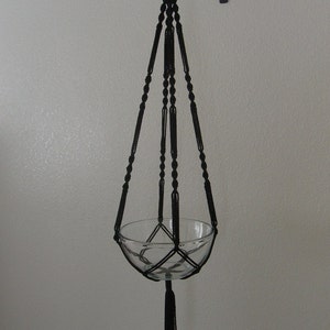 May include: Black macrame plant hanger with a clear glass bowl. The hanger is suspended from a decorative black metal hook attached to a white wall. The macrame design features intricate knots and a tassel at the bottom.