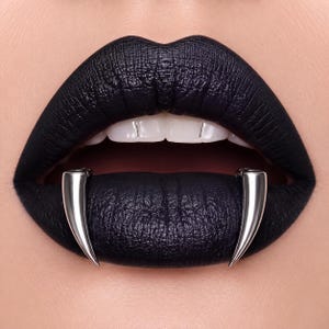 May include: A pair of polished silver-toned metal fangs worn over the lower lip, contrasting against matte black lipstick. The fangs are curved, pointed, and metallic, positioned symmetrically on the lower lip with white teeth visible above.
