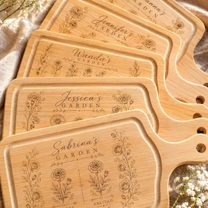 May include: A stack of personalized wooden cutting boards with floral designs and names like "Adrianna's Garden" and "Sabrina's Garden" engraved on them. The boards have a rectangular shape with a handle and are made of light-colored wood.