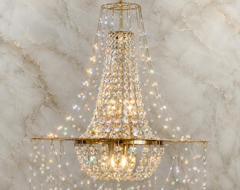 May include: A gold-toned chandelier with multiple tiers of sparkling crystals. The fixture has a central, bell-shaped design with a series of hanging crystal strands. The light fixture is suspended from a gold chain and is set against a marble-like background.