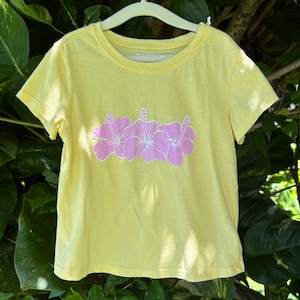 May include: A bright yellow short-sleeve t-shirt featuring a graphic print of three light pink hibiscus flowers centered on the chest. The shirt has a round neckline and is displayed on a light-colored hanger against a background of green leaves.