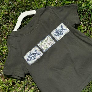 May include: A dark olive green short-sleeve t-shirt featuring three rectangular patches arranged diagonally across the chest. Each patch displays a stylized fish graphic with intricate geometric patterns in shades of blue, green, and white.