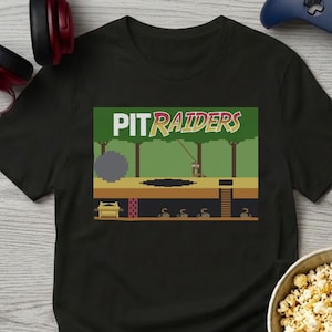 May include: A black short-sleeve t-shirt featuring a pixelated graphic print of a video game scene with the text PIT RAIDERS in white and yellow letters. The design depicts a jungle landscape, a boulder, a pit, and an underground treasure chamber.