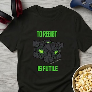 May include: A black short-sleeved t-shirt featuring a graphic of a dark gray mechanical cube with glowing green heart symbols on its faces. The text "TO RESIST IS FUTILE" is printed in bright green above and below the cube illustration.