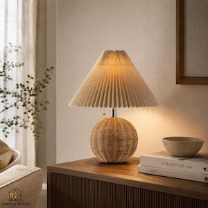Op de afbeelding: Een tafellamp met een beige geplooide lampenkap en een geweven rotan voet. De lamp staat op een houten oppervlak, met een kom en boeken. Het ontwerp heeft natuurlijke tinten en texturen.