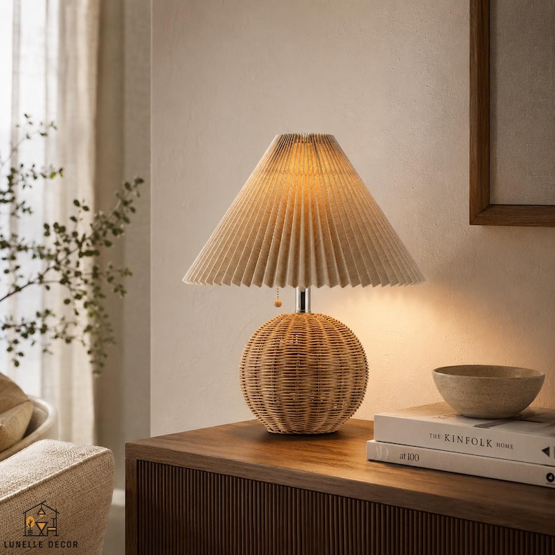 Op de afbeelding: Een tafellamp met een beige geplooide lampenkap en een geweven rotan voet. De lamp staat op een houten oppervlak, met een kom en boeken. Het ontwerp heeft natuurlijke tinten en texturen.