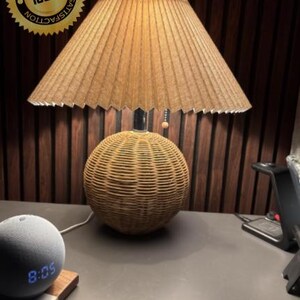 Op de afbeelding: Een decoratieve tafellamp met een geplooide lampenkap en een geweven rotan voet. De lamp is bruin en beige, met een ronde voet en een conische kap. De tijd 8:05 wordt weergegeven op een grijze slimme luidspreker.