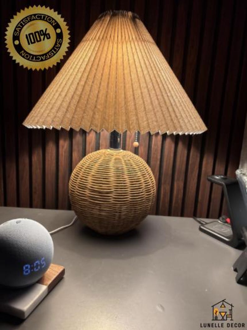 Op de afbeelding: Een decoratieve tafellamp met een geplooide lampenkap en een geweven rotan voet. De lamp is bruin en beige, met een ronde voet en een conische kap. De tijd 8:05 wordt weergegeven op een grijze slimme luidspreker.