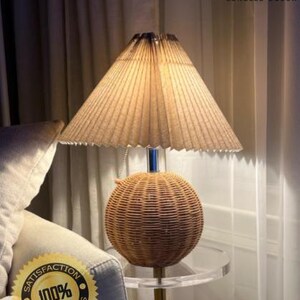 Op de afbeelding: Een decoratieve tafellamp met een basis van geweven rotan en een geplooide lampenkap. De lamp staat op een transparante acryl bijzettafel met een goudkleurige metalen standaard. De lamp is verlicht en geeft een warme gloed. De tekst "LUNELLE DECOR" en een "100% Satisfaction" sticker zijn inbegrepen.