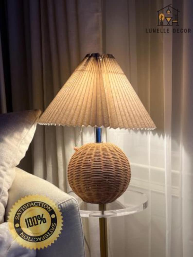 Op de afbeelding: Een decoratieve tafellamp met een basis van geweven rotan en een geplooide lampenkap. De lamp staat op een transparante acryl bijzettafel met een goudkleurige metalen standaard. De lamp is verlicht en geeft een warme gloed. De tekst "LUNELLE DECOR" en een "100% Satisfaction" sticker zijn inbegrepen.