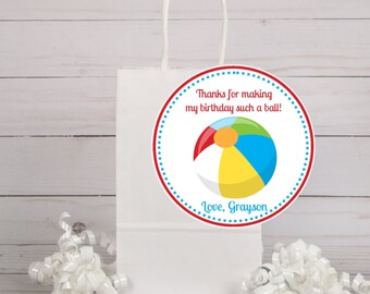May include: White paper gift bag with a round sticker featuring a colorful beach ball and the text "Thanks for making my birthday such a ball! Love, Grayson".