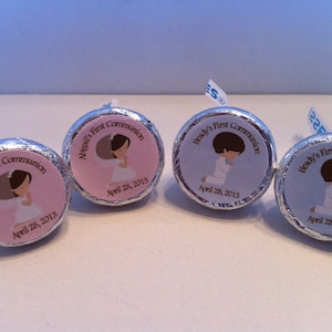 May include: Four round, silver foil-wrapped chocolate candies with personalized labels. The labels feature a pink, blue, or white background with a cartoon image of a child in a white dress or suit praying. The labels also include the text "First Communion" and the child's name, along with the date "April 28, 2013".