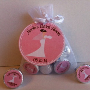 May include: Pink and white bridal shower favor bags filled with chocolate kisses. The bags have a pink circle label with a bride in a white dress and the text "Nicole's Bridal Shower 05.15.14". The chocolate kisses have a smaller pink label with a bride and groom and the text "It all started with a kiss Nicole's Bridal Shower 05.15.14".