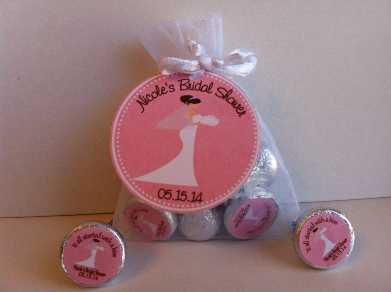 May include: Pink and white bridal shower favor bags filled with chocolate kisses. The bags have a pink circle label with a bride in a white dress and the text "Nicole's Bridal Shower 05.15.14". The chocolate kisses have a smaller pink label with a bride and groom and the text "It all started with a kiss Nicole's Bridal Shower 05.15.14".