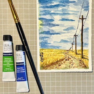 Pode incluir: Uma aguarela de uma paisagem rural com um caminho de terra, campos amarelos e postes de eletricidade sob um céu azul com nuvens, acompanhada por um pincel de cabo preto e dois tubos de aguarela Winsor & Newton de 10 ml em verde e azul.